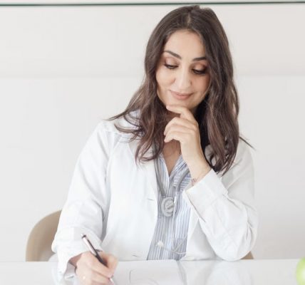 A female nutritionist in a lab coat writing notes with an apple and plant on the desk.